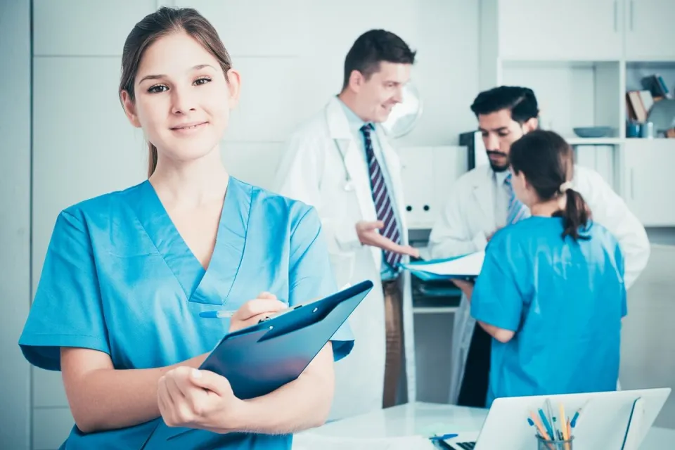 Smiling Medical Assistant with Clipboard with Doctors in Background